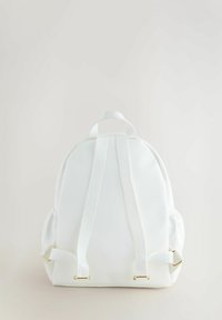 Sac à dos blanc en matériau texturé, avec des bretelles rembourrées, une forme ronde et une subtile marque sur les bretelles. Accents en métal doré.