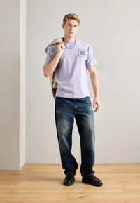 Lys lilla t-skjorte med korte ermer og grafisk trykk, kombinert med falmede blå rette jeans og svarte kraftige sko. Beige jakke over skulderen.