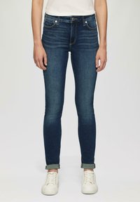 Skinny denim jeans in dark blue with a smooth texture, classic five-pocket design, and rolled cuffs. Worn with white sneakers.
