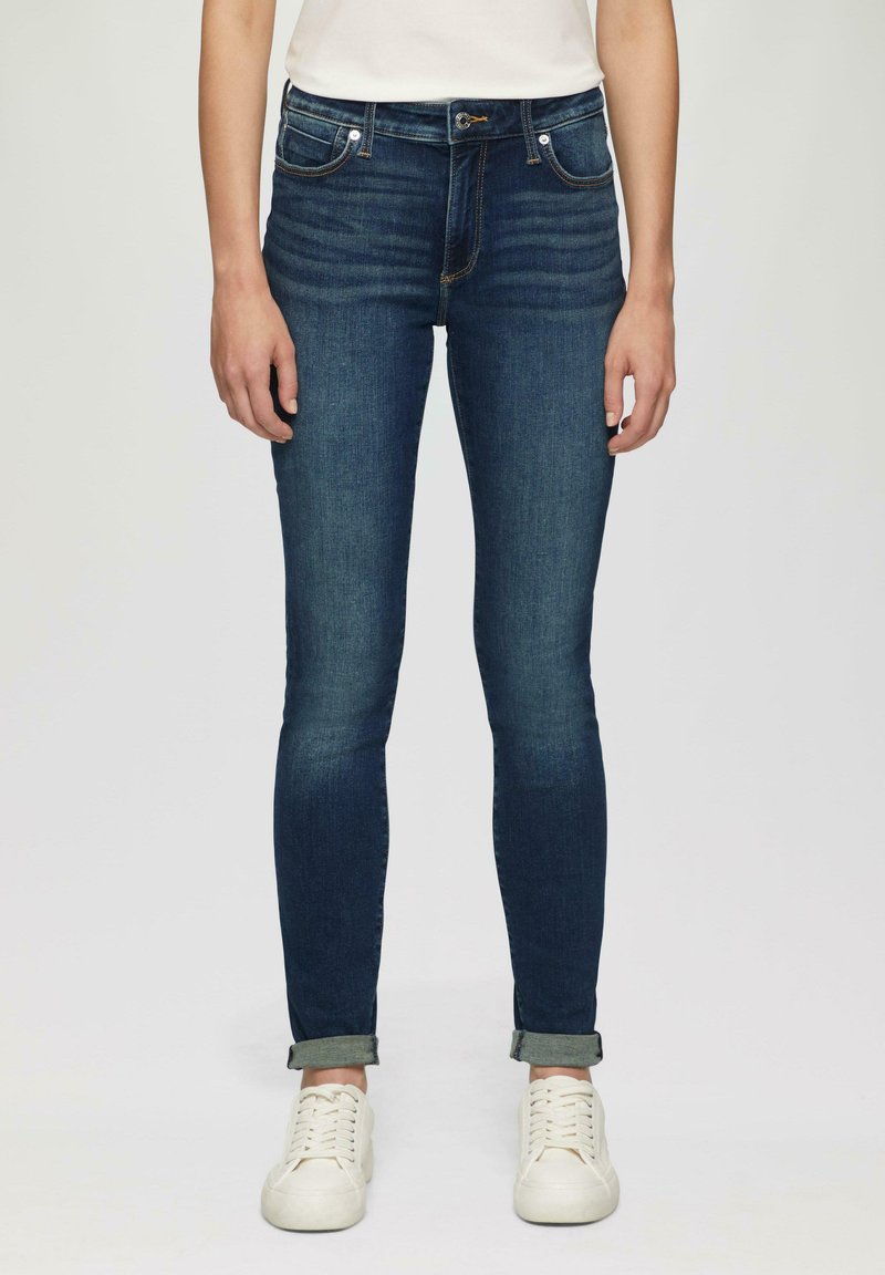 Skinny denim jeans in dark blue with a smooth texture, classic five-pocket design, and rolled cuffs. Worn with white sneakers.