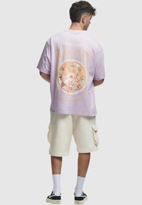 Lilla oversized t-skjorte med en fargerik sirkulær grafikk på baksiden, kombinert med kremfargede cargo-shorts og svarte sneakers.