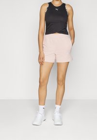 Top nero senza maniche con un design attorcigliato, abbinato a pantaloni corti rosa chiaro con tasche laterali. Sneakers bianche e calze completano l'outfit.
