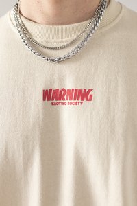 Beige Baumwoll-T-Shirt mit rotem "WARNUNG"-Text und "KAOTIKO SOCIETY"-Druck. Rundhalsausschnitt, kurze Ärmel, lockere Passform. Mehrere silberne Ketten.
