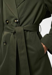 Topeltkarvaline tumeroheline mantel, millel on seotud vöökinnitus, kilpkonnamustriga nööbid ja käsi paremas taskus, neutraalse taustaga.