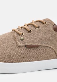 Brown fabric sneaker with light brown laces, gold eyelets, and a tan leather accent. Features a white rubber sole and textured fabric.