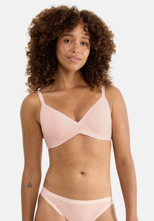 Frau mit lockigem Haar und Sommersprossen, die einen hellrosa gerippten BH und passende Slips trägt, vor einem schlichten weißen Hintergrund stehend.