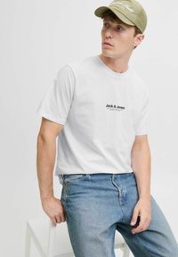 Weißes Baumwoll-T-Shirt mit schwarzem "Jack & Jones"-Logo auf der Brust, kombiniert mit hellblauen Jeans. Das Modell trägt eine beige Mütze.