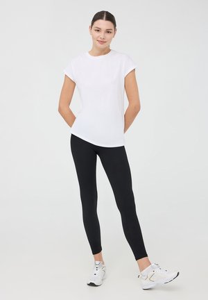 Biała koszulka z krótkim rękawem zestawiona z czarnymi legginsami. Model nosi białe buty sportowe. Gładki materiał o dopasowanym kroju.