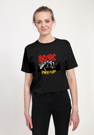Černé bavlněné tričko s odvážným grafickým potiskem, na kterém je v červené barvě nápis "AC/DC" a v žluté barvě "PWR UP", spolu s monochromatickým obrázkem kapely.