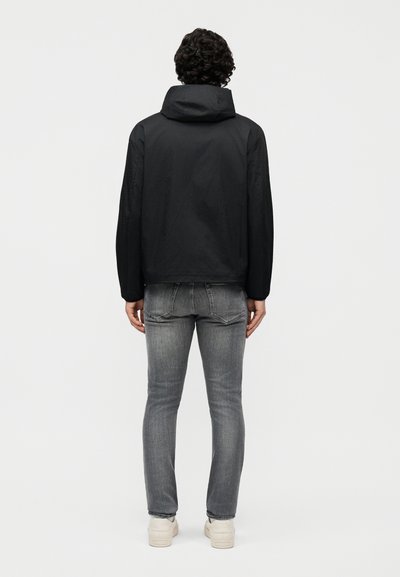 Göndör hajú férfi fekete kapucnis dzsekiben, szürke farmerral és fehér tornacipővel, háttal áll egy egyszínű háttér előtt.