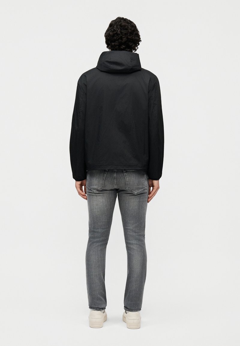 Uomo con capelli ricci che indossa una giacca nera con cappuccio, jeans grigi e sneakers bianche, in piedi di schiena contro uno sfondo neutro.