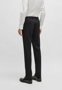 Pantaloni neri dal design sartoriale con una texture liscia. Presentano tasche posteriori doppie e passanti per cintura. Indossati con una camicia bianca e scarpe nere.