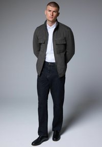 Veste en suède gris avec deux poches poitrine, chemise blanche à boutons, pantalon en denim sombre et chaussures en cuir noires. Le mannequin se tient face à l'avant.