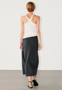 Débardeur blanc à larges bretelles, associé à une longue jupe gris foncé. Sandales noires à lacets autour de la cheville complètent la tenue. Design minimaliste.