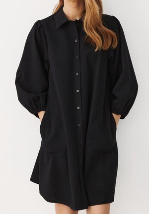 Kvinde iført en sort tekstureret knap-op kjole med trekvart lange puff ærmer og hænderne i sidelommer.