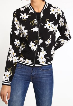 Woman wearing a black zip-up jacket with white floral print and striped cuffs, paired with blue denim jeans.