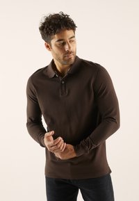 Polo à manches longues marron en tissu doux. Présente un col classique, une patte de boutonnage à un seul bouton et un design minimaliste sans motifs.