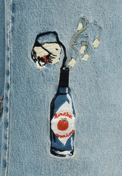 Το μπλε τζιν ύφασμα διαθέτει κεντημένη εικόνα ενός μπουκαλιού με κόκκινη ετικέτα, κορυφωμένο με στυλιζαρισμένη εκτόξευση και διακοσμημένο με λευκές λεπτομέρειες.