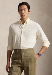 Mann trägt cremefarbenes Hemd mit Knöpfen, hochgeschnittene olivgrüne Hose und eine Armbanduhr mit blauem Zifferblatt und braunem Lederarmband.