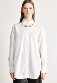 Λευκό πουκάμισο με μακριά μανίκια, κουμπιά μπροστά, απαλή υφή και τσέπη στο στήθος. Συνδυασμένο με μεταλλική αλυσίδα και μαύρα παντελόνια με υφή.