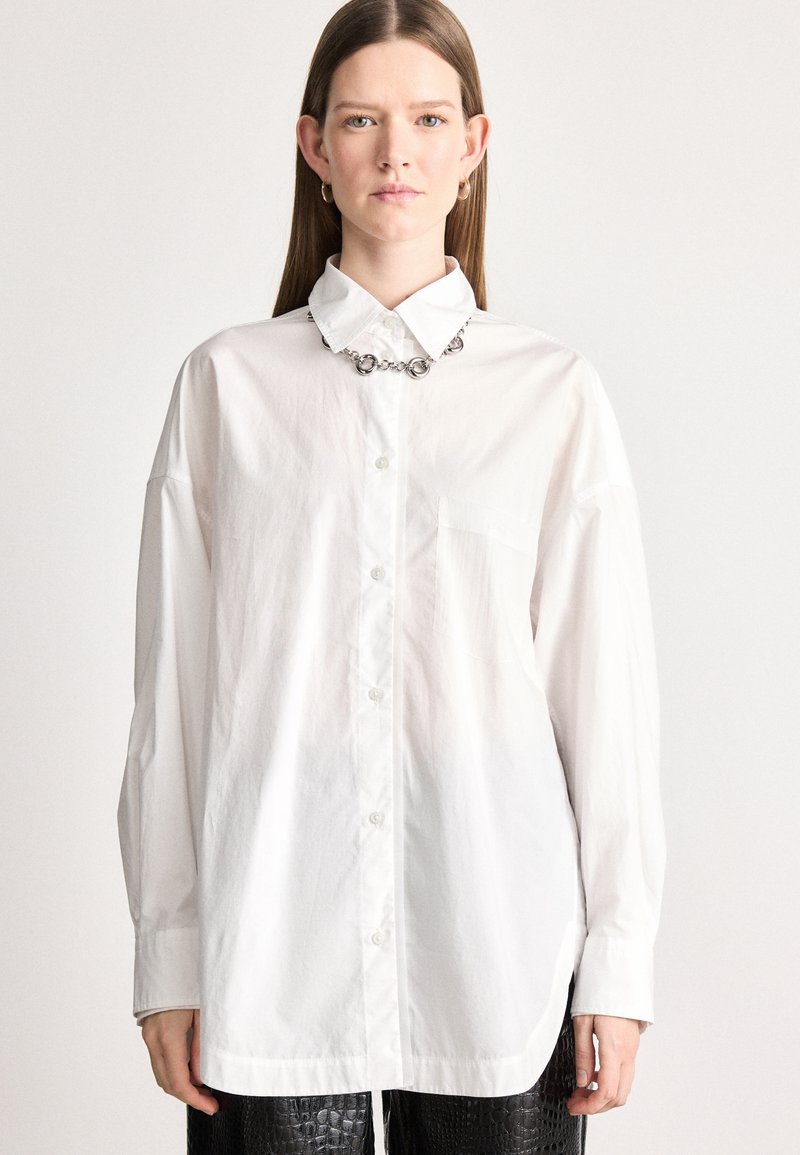 Λευκό πουκάμισο με μακριά μανίκια, κουμπιά μπροστά, απαλή υφή και τσέπη στο στήθος. Συνδυασμένο με μεταλλική αλυσίδα και μαύρα παντελόνια με υφή.