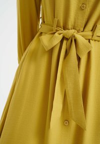 Abito giallo a camicia con tessuto testurizzato, dotato di una cintura annodata in vita e una silhouette fluida. I bottoni sono piccoli e rotondi.