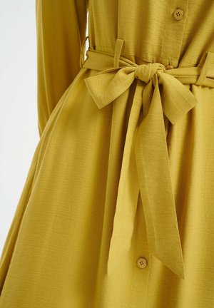 Gelbes Hemdkleid aus strukturiertem Stoff, mit einem gebundenen Taillengürtel und einer fließenden Silhouette. Die Knöpfe sind klein und rund.