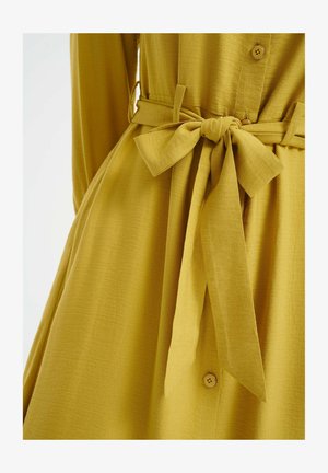 Geel knoopjurk met een gestructureerde stof, voorzien van een gestrikte tailleband en een vloeiende silhouet. De knopen zijn klein en rond.