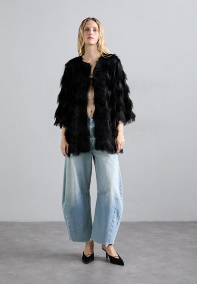 Melnā pūkainā mākslīgā kažokā, plata gaismas zila džinsu bikses ar saplēstu malu un melni apavi ar norādītu purngalu. Minimālistisks fons.