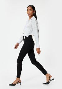 Camisa blanca corta con bolsillo en el frente, combinada con pantalones negros ajustados. La modelo lleva tacones slingback de charol negros. Diseño simple y limpio.