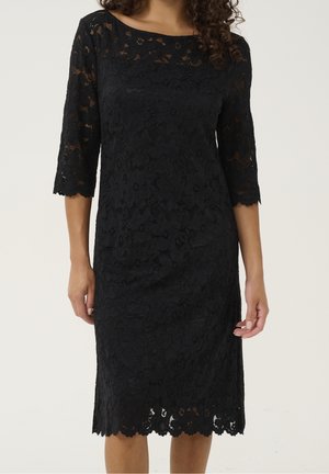 Frau trägt ein knieumspieltes schwarzes Spitzenkleid mit dreiviertellangen Ärmeln und wellenförmigen Kanten an Ärmeln und Saum.