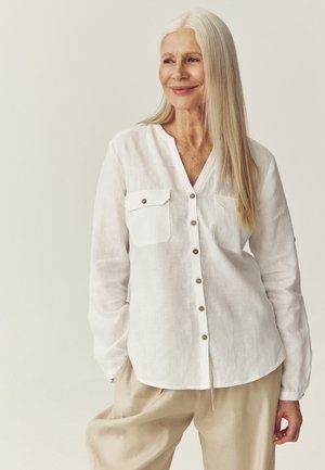 Uśmiechnięta starsza kobieta z długimi siwymi włosami, ubrana w białą, zapinaną na guziki koszulę i beżowe spodnie, stojąca na jasnym, jednolitym tle.