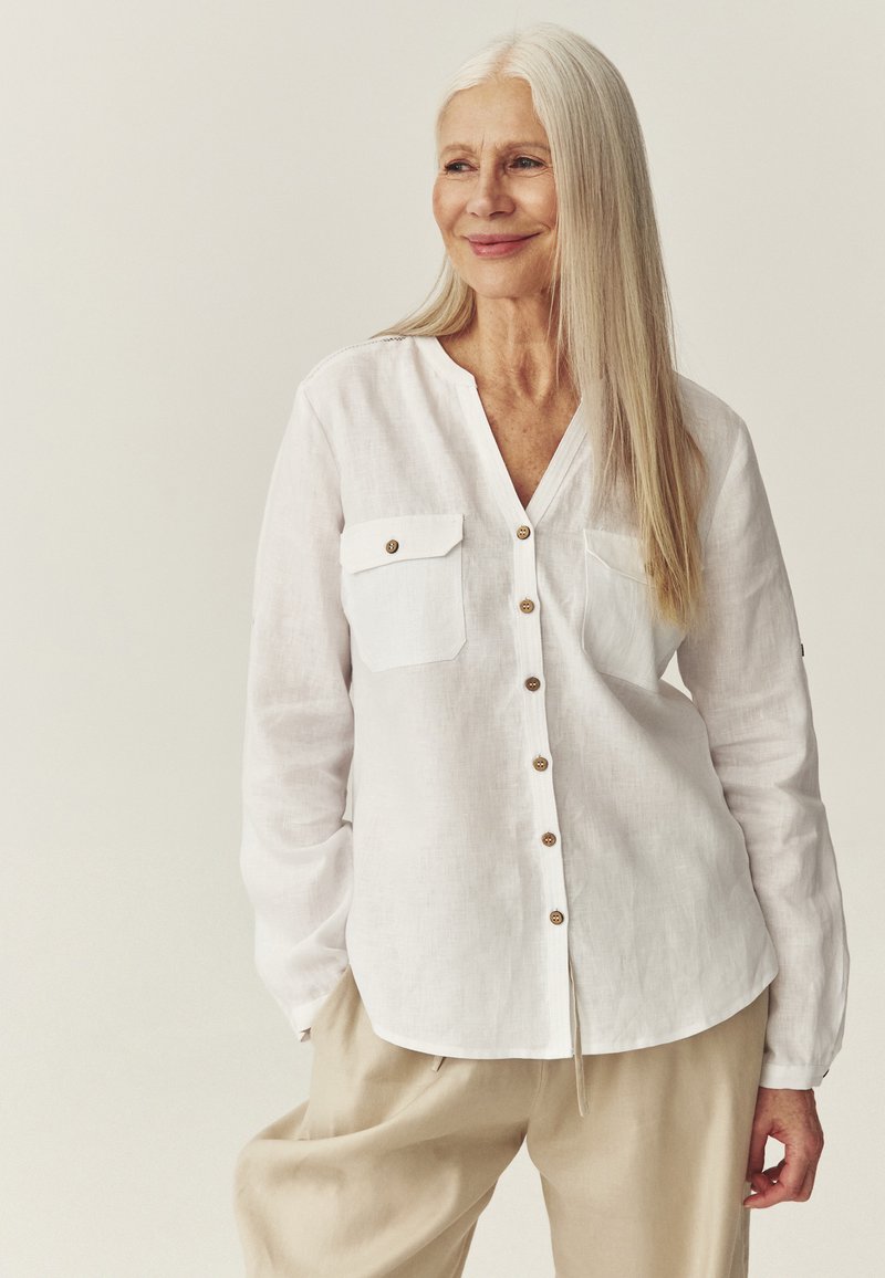 Femme âgée souriante aux longs cheveux gris, portant une chemise blanche boutonnée et un pantalon beige, debout devant un fond clair uni.
