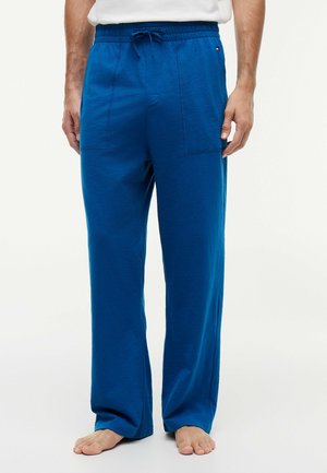 Mann trägt locker sitzende blaue Hose mit Kordelzug und weißes Hemd, steht barfuß vor hellem, einfarbigem Hintergrund.