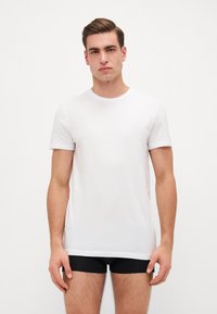 T-shirt blanche à manches courtes en tissu doux, encolure ronde, coupe slim, portée avec un short noir, présentée sur un fond lumineux.