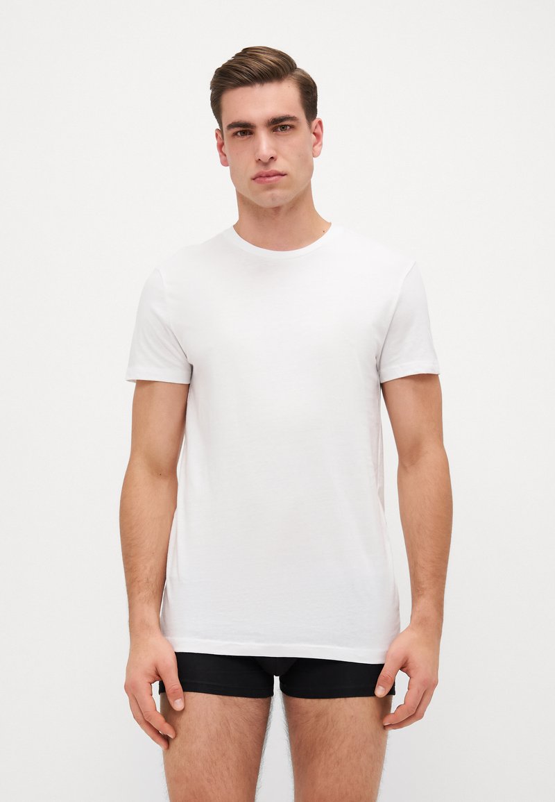 Schlichtes weißes T-Shirt mit kurzen Ärmeln aus weichem Stoff, runder Ausschnitt, schlanker Schnitt, getragen mit schwarzen Shorts, vor einem hellen Hintergrund präsentiert.