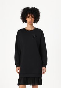 Robe en sweatshirt noire avec des manches allongées, un ourlet côtelé et une jupe plissée. Accentuée d'un logo orné de perles. Texture de tissu douce.