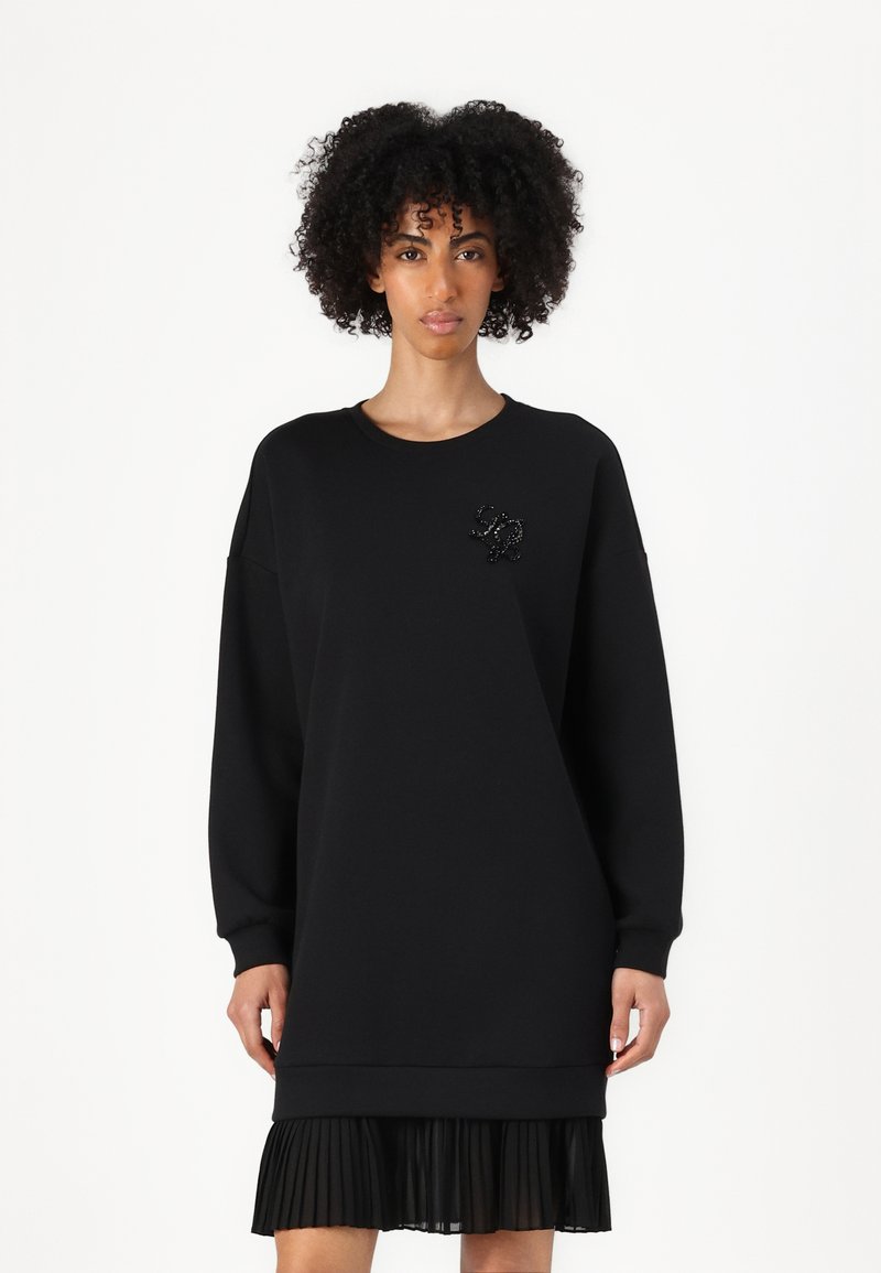 Robe en sweatshirt noire avec des manches allongées, un ourlet côtelé et une jupe plissée. Accentuée d'un logo orné de perles. Texture de tissu douce.