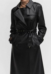 Femme portant un trench-coat en cuir noir ceinturé, avec des détails de boutons et les mains dans les poches, montrée du cou jusqu'au milieu de la cuisse.