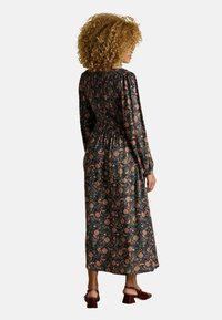 Langes, dunkles Blumenkleid mit langen Ärmeln und einem gerafften Rücken. Hat ein farbenfrohes Muster aus Rosen auf schwarzem Hintergrund. Wird mit Absatzsandalen getragen.