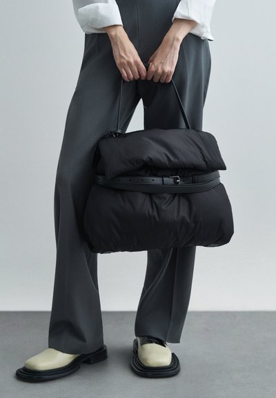 Μαύρη μαλακή τσάντα με απλή υφή, που διαθέτει λουρί σαν ζώνη. Φορεμένη με γκρι κομμένα παντελόνια και μπεζ παπούτσια με μαύρες σόλες.