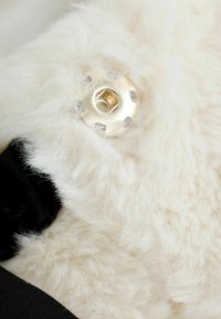 Pūkuota grietinėlės spalvos tekstilė su auksiniu metaliniu tvirtinimu. Fone matoma juoda medžiaga, akcentuojanti kontrastingas tekstūras ir spalvas.