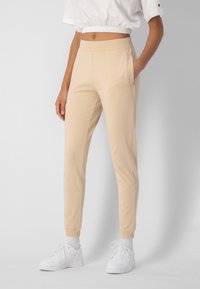 Beige Jogginghosen aus weichem Stoff, mit einem taillierten Bund, Seitentaschen und gerippten Bündchen. Kombiniert mit weißen Sportschuhen.