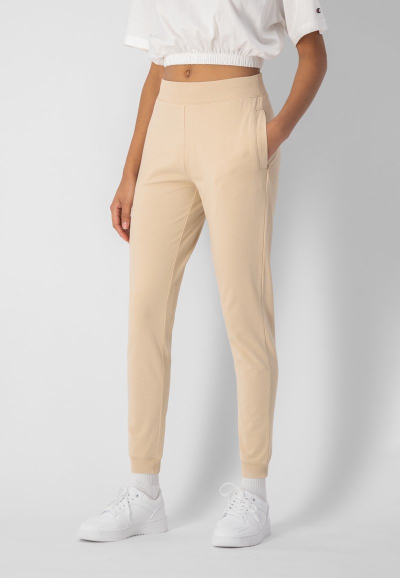 Beige Jogginghosen aus weichem Stoff, mit einem taillierten Bund, Seitentaschen und gerippten Bündchen. Kombiniert mit weißen Sportschuhen.