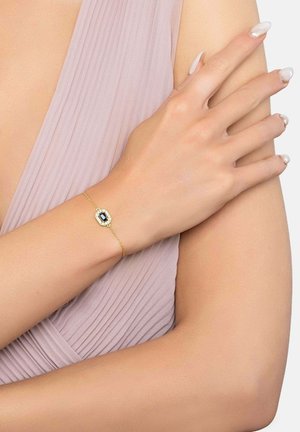 Delicaat gouden armband met een rechthoekige donkerblauwe edelsteen en kleine diamanten, gedragen om de pols tegen lichtroze geplooide stof.