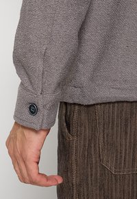 Grijs textuur langemouwen shirt met knoopmanchet details, gecombineerd met bruine pantalon van gestreepte stof. Focus op stofoppervlak en pasvorm.