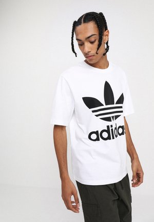 T-shirt bianco di cotone con un grande logo Adidas nero e un design a foglia. Maniche corte e collo rotondo. Indossato con pantaloni verde scuro.
