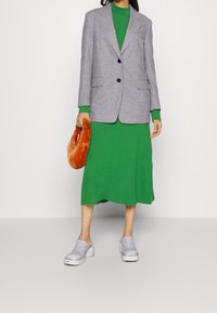 Blazer gris à deux boutons porté sur une robe en tricot vert. Des chaussures grises sans lacets et un sac à main en peluche orange complètent la tenue.