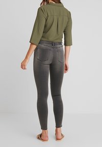 Mujer con blusa verde oliva de manga tres cuartos, jeans ajustados grises y sandalias planas con estampado de cebra, de pie de espaldas sobre fondo blanco.