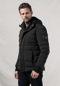 Giacca a vento nera con cappuccio, caratterizzata da un design trapuntato, tasche laterali e chiusura con zip frontale. Realizzata in un materiale liscio e durevole.